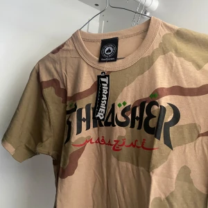 Beige camo t-shirt från Thrasher - Säljer en beige och grön camo t-shirt från Thrasher med tryck på bröstet. T-shirten har rund halsringning och korta ärmar. Perfekt för dig som gillar streetwear och vill sticka ut lite extra.