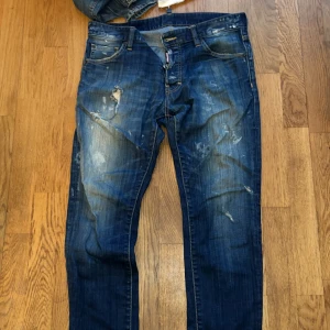 Blå jeansbyxor från Dsquared2, Nypris 6000kr - Säljer ett par blå jeansbyxor från Dsquared2 med slitna detaljer och ljusa tvättade partier. Byxorna har klassisk femficksdesign, knappgylf och Dsquared2-logga vid fickan. Perfekta för dig som gillar en trendig och avslappnad stil.