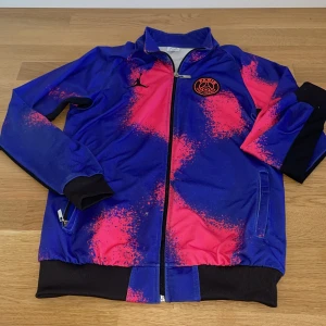 PSG x Air Jordan träningsjacka blå/rosa - Säljer en snygg PSG x Air Jordan träningsjacka i blått och rosa med svart logga och Paris Saint-Germain-märke på bröstet. Jackan har dragkedja, ribbade muddar och coolt färgstänksmönster. Perfekt för dig som gillar fotboll och streetwear.