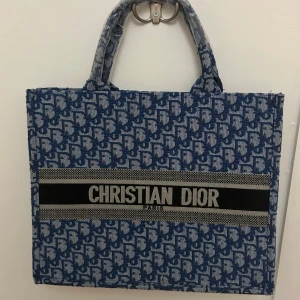 Blå shoppingväska från Christian Dior - Säljer en ikonisk blå shoppingväska från Christian Dior med tydligt logomönster och breda handtag. Väskan har ett stort fack och är dekorerad med märkets namn på framsidan. Perfekt för dig som vill 