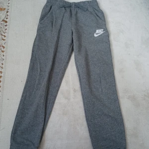 Grå mjukisbyxor från Nike - Grå mjukisbyxor från Nike med resår i midjan och raka ben. Byxorna har en vit Nike-logga broderad på ena benet och är tillverkade i mjuk bomull. Perfekta för en avslappnad stil.