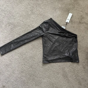 Glittrig oneshoulder topp från Noisy May - Säljer en svartglittrig oneshoulder topp från Noisy May. Toppen har en lång ärm och sitter tight. Perfekt för dig som vill sticka ut med en snygg och trendig look.