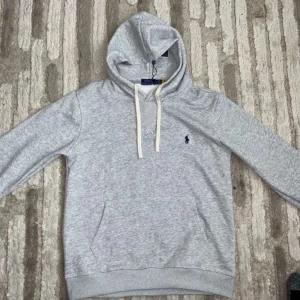 Grå hoodie från Polo Ralph Lauren - Säljer en klassisk grå hoodie från Polo Ralph Lauren med broderad logga på bröstet. Tröjan har huva med vita snören och en stor ficka framtill. Perfekt för en avslappnad stil.