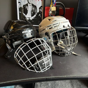 Bauer hockeyhjälmar svart och vit - Två hockeyhjälmar från Bauer, en svart och en vit, båda med galler för ansiktsskydd. Klassisk rundad form och justerbar passform. Perfekt för dig som spelar hockey och vill ha bra skydd och stilren design.