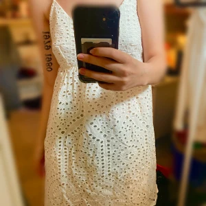 Vit broderad sommarklänning med spetsdetaljer - Söt vit klänning med broderade spetsdetaljer och smala axelband. Djup v-ringning och knyt i ryggen. Perfekt för varma dagar och midsommarfirande. Kort modell med vågig kant nertill och luftig passform. Aldrig använd.