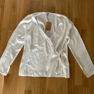 Vit blus från NA-KD - Säljer en vit blus från NA-KD med långa ärmar och knyt i sidan. Köpt från sellpy och har endast testat den. Kort i armarna!