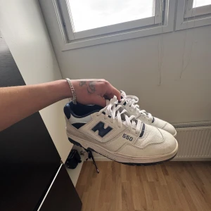 New Balance 550 – White/Navy, EU44.5 (Retro Basketstil) - Säljer ett par New Balance 550 sneakers i vitt och marinblått. Klassisk retrodesign med tydlig New Balance-logga på sidan och hälen. Skorna har snörning och platt sula, perfekta för en sportig och stilren look.