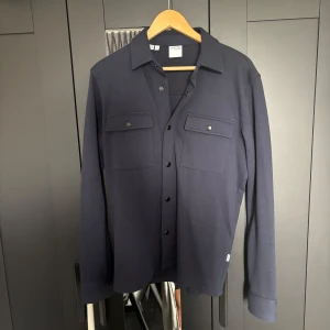 Mörkblå overshirt från Selected Homme - Snygg mörkblå overshirt från Selected Homme med klassisk krage och två stora bröstfickor med knappar. Skjortan har långa ärmar och stängs med svarta knappar framtill. Perfekt att bära som lager över en t-shirt.