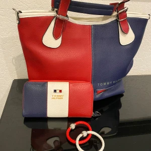 Flerfärgad handväska och plånbok från Tommy Hilfiger, använd endast 2 gånger - Säljer en snygg handväska och matchande plånbok från Tommy Hilfiger i rött, blått och vitt. Väskan har två handtag och är tillverkad i syntetmaterial med tydlig logga. Plånboken har samma färgblock och logotyp framtill. Har även 2 matchande armband.