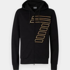 Svart hoodie EA7 med guldiga text - Svart hoodie från EA7 Emporio Armani med dragkedja och huva. Framsidan har ett stort gult grafiskt tryck i form av en sjua. Liten EA7-logga i gult på ryggen nedtill. Perfekt för en sportig och stilren look.