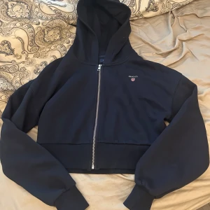 Marin blå croppad hoodie från Gant - Säljer en marinblå croppad hoodie från Gant med dragkedja framtill och liten logga på bröstet. Tröjan har långa ärmar och en stor huva. Perfekt för en avslappnad och trendig stil. Storlek 170 men funkar som xs/s