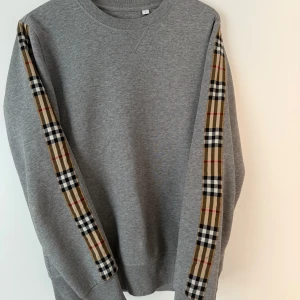 Grå sweatshirt från Burberry med rutiga detaljer - Säljer en grå sweatshirt från Burberry med klassiska beige, svarta och röda rutiga band längs ärmarna. Tröjan har rund halsringning och är tillverkad i mjuk bomullsblandning.