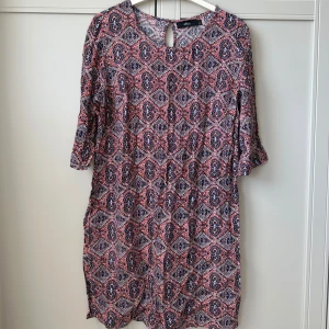 Mönstrad klänning från Ellos - Kortärmad klänning från Ellos med färgstarkt mönster i rosa, blått och vitt. Klänningen har en rak passform, rund halsringning och knappdetalj i nacken. Perfekt för dig som gillar boho-vibbar och vill ha något bekvämt men snyggt.