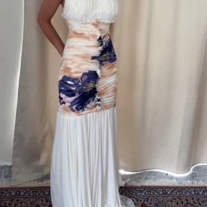 Vit och mönstrad långklänning med öppen rygg - Säljer en elegant långklänning i vitt med abstrakt mönster i lila, beige och blått. Klänningen har plisserad överdel, smala axelband och öppen rygg med snygga banddetaljer. Kjolen är lång och faller mjukt. Perfekt för speciella tillfällen! Köpt från USA på 15000kr. Haft på mig en gång bara 