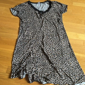 Leopardmönstrad kortklänning med spetsdetalj - Kortklänning med leopardmönster och v-ringning med svart spetskant. Klänningen har korta ärmar och är tillverkad i ett mjukt, lätt material. Perfekt för dig som gillar djurmönster och vill sticka ut.