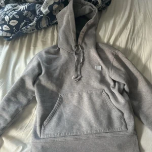Grå hoodie från Acne Studios - Säljer en stilren grå hoodie från Acne Studios med klassisk smiley-patch på bröstet. Tröjan har huva med dragsko och stor magficka framtill. Perfekt för en avslappnad och trendig look. Lite slit på ärmena men inget man riktigt märker samt kan den göra så tröjan blir lite snyggare