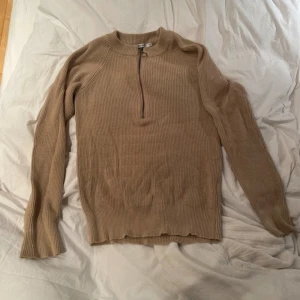 Beige stickad tröja med dragkedja från NA-KD - Säljer en beige stickad tröja från NA-KD i storlek XS. Tröjan har lång ärm, hög krage och en snygg dragkedja framtill med rund detalj. Perfekt för lager-på-lager och enkel att matcha med olika outfits. Den ser ganska skrynklig ut på bilden bara 🙂‍↕️