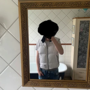 Vit dunväst från J.Lindeberg - Säljer en stilren vit dunväst från J.Lindeberg med dragkedja framtill och två fickor med dragkedja. Västen har en quiltad design och hög krage. Perfekt för lager-på-lager och enkel att matcha med olika outfits.