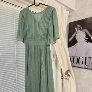 Ljusgrön plisserad långklänning - Säljer en elegant ljusgrön långklänning med plisserat tyg och lös passform. Klänningen har korta, vida ärmar och markerad midja. Perfekt för dig som vill ha en stilren och luftig look.