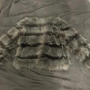 Grå pälsjacka med ränder - Säljer en fluffig  grå pälsjacka med mörk grå ränder. Jackan har lång ärm och en boxig, lite oversized passform. Perfekt för dig som vill sticka ut med en trendig look. Jackan är mjuk och har en lyxig känsla.