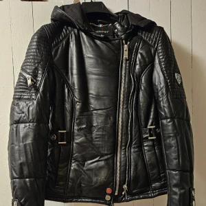 Svart dun skinnjacka med huva från Horspist - Säljer en svart skinnjacka från Horspist med huva och quiltade detaljer på axlar och ärmar. Jackan har dragkedja framtill, spännen i midjan och flera snygga sömmar som ger en cool look. Jackan är väldigt liten i storleken/ motsvarar storlek XS. Finns också stor päls till luvan om man önskar det.