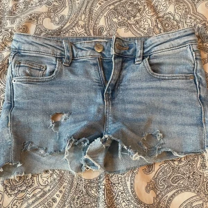 Ljusblå jeansshorts med fransar från Gina Tricot - Säljer ett par ljusblå jeansshorts från Gina Tricot med slitna detaljer och fransig kant. Klassisk femficksmodell med knapp och dragkedja framtill. Perfekta för sommaren! Storlek 140 i barnstorlek