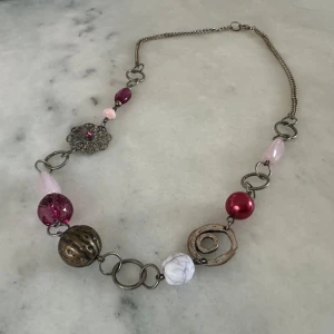 Färgglatt halsband med stora pärlor och metallblommor - Unikt halsband med stora pärlor i rosa, vitt och brons samt dekorativa metallblommor och länkar. Kedjan är i silverfärgad metall och halsbandet har en mix av former och färger som ger ett lekfullt och trendigt intryck.