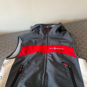 Grå och röd väst från Sail Racing - Snygg grå väst från Sail Racing med röda detaljer och dragkedjor. Västen har en huva och är perfekt för utomhusaktiviteter. Den har en sportig look med logotyp på bröstet och sidan.
