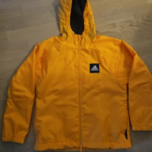 Gul vindjacka från Adidas - Säljer en snygg orangel vindjacka från Adidas med huva och dragkedja. Jackan har en svart logotyp på bröstet och är perfekt för blåsiga dagar. Den har långa ärmar och en bekväm passform.
