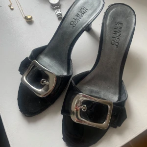 Svarta sandaler med silverdetalj från Franco Sarto - Snygga svarta sandaler från Franco Sarto med en stor silverfärgad spänne på ovansidan. De har en öppen tå och en elegant klack som ger en stilren look. Skulle säga att de är i storlek 38, 40 och lätta att gå i.