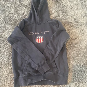 Mörkblå hoodie från GANT - Säljer en snygg mörkblå hoodie från GANT som inte säljs längre där jag köpte för 500 med ett coolt emblem på bröstet. Tröjan har en klassisk känguruficka och en bekväm huva. Perfekt för en avslappnad stil. Passar ungdomar i åldern 13-14 år.