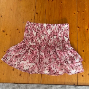 Rosa blommig kjol från Gina - Charmig rosa blommig kjol med volangdetaljer. Perfekt för sommarens varma dagar. Den har en elastisk midja för bekväm passform.köpt på Gina och är använd två gånger