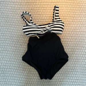 Randig bikini i svart och vit - Snygg baddräkt med svart och vit randigt mönster på toppen och enfärgad svart underdel. Toppen har breda axelband och en klassisk skärning. Perfekt för en dag på stranden eller vid poolen.