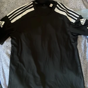 Svart t-shirt från Adidas - Säljer en svart t-shirt från Adidas med de klassiska tre vita ränderna på axlarna. T-shirten har korta ärmar och är tillverkad i ett syntetmaterial som är perfekt för träning. 