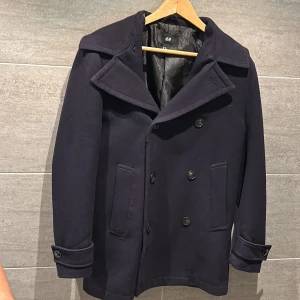 Mörkblå skepparkavaj från H&M - Säljer en stilren mörkblå skepparkavaj från H&M. Jackan har en klassisk design med bred krage och knappar framtill. Perfekt för kyligare dagar med sin långa ärm och eleganta look. Passar både till vardag och mer uppklädda tillfällen.