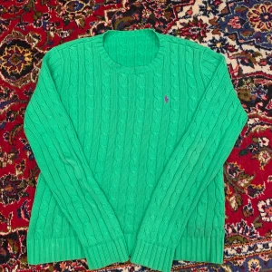 Grön kabelstickad tröja från Ralph Lauren - Snygg grön kabelstickad tröja från Ralph Lauren med lila broderad logga på bröstet. Tröjan har en klassisk rund halsringning och långa ärmar. Perfekt för en stilren look!