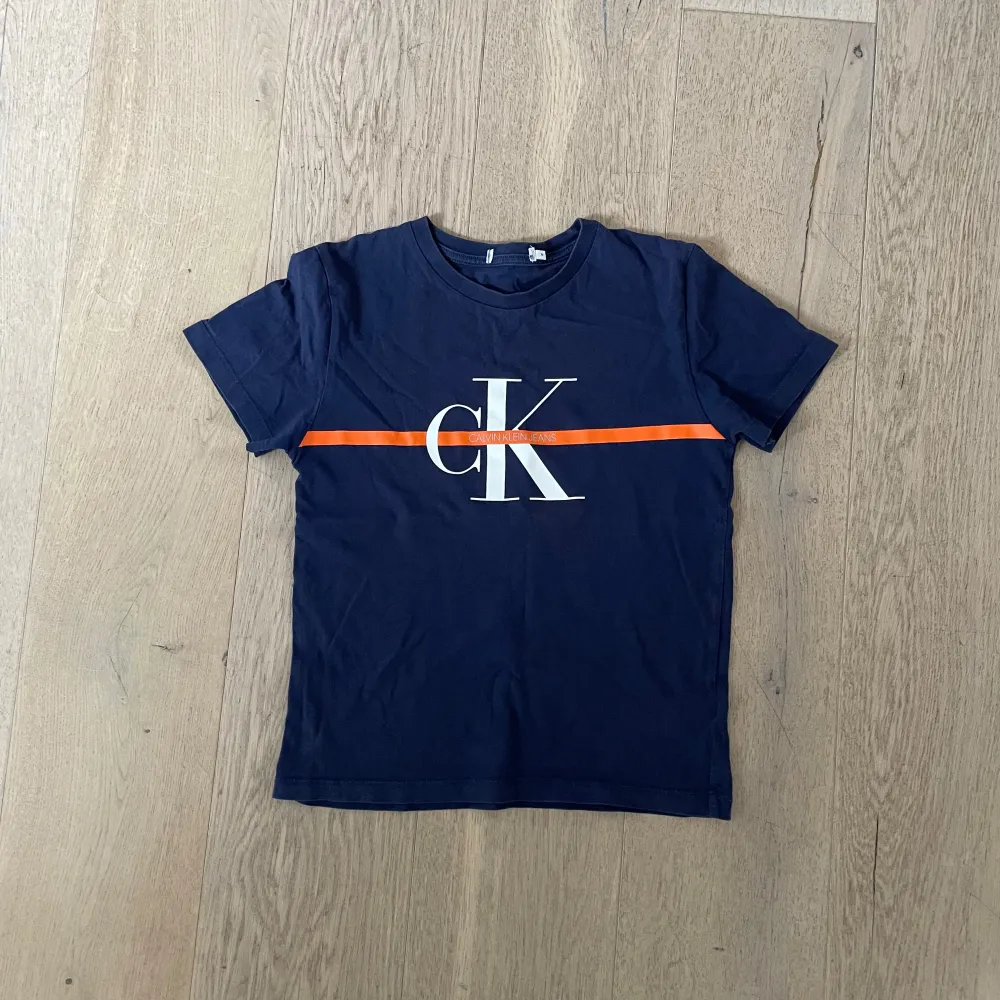 Snygg mörkblå t-shirt från Calvin Klein med en orange rand och CK-logga på bröstet. Perfekt för en stilren look.. T-paidat.
