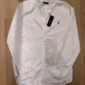 Vit skjorta från Ralph Lauren - Säljer en klassisk vit skjorta från Ralph Lauren med broderad logga på bröstet. Skjortan har långa ärmar och knappar framtill. Perfekt för en stilren look.