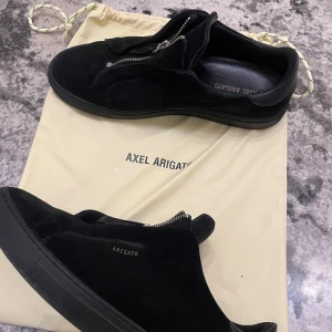 Svarta sneakers från Axel Arigato - Snygga svarta sneakers i mocka från Axel Arigato med dragkedja framtill. Perfekta för en stilren look. Skorna kommer med en praktisk dustbag för förvaring. Och låda 