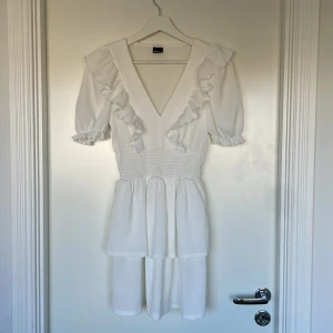 Vit klänning från Gina Tricot - Säljer en söt vit klänning från Gina Tricot med volangdetaljer och v-ringning. Klänningen har korta ärmar och en smickrande passform. Perfekt för sommaren eller skolavslutning! Bara använd 1 gång, inga fläckar eller hål, som ny!💝