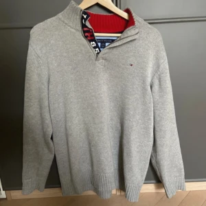 Grå tröja med dragkedja från Tommy Hilfiger - Säljer en stilren grå tröja från Tommy Hilfiger med dragkedja och ribbad krage. Tröjan har långa ärmar och mörkblå armbågslappar som ger en snygg kontrast. Perfekt för en avslappnad och trendig look.