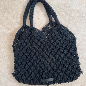 Svart stickad väska från Don Donna - Snygg svart stickad väska med ett flätat mönster. Perfekt för en dag på stranden eller för att handla på marknaden. Väskan har två handtag och en öppen design 🖤