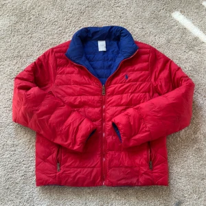 Vändbar jacka från Ralph Lauren - Säljer en snygg vändbar pufferjacka från Ralph Lauren. Ena sidan är röd och den andra blå, båda med dragkedja och broderad logga på bröstet. Perfekt för att variera stilen beroende på. Jackan har långa ärmar och är quiltad för extra värme.