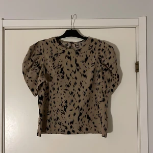 Beige blus med svart mönster - Snygg beige blus med svart mönster och puffiga korta ärmar. Perfekt för en stilren look. Blusen har en rund halsringning och en knappdetalj i nacken. Oanvänd!