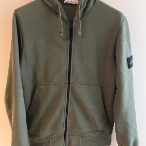 Grön hoodie från Stone Island - Säljer en snygg grön hoodie från Stone Island med dragkedja och justerbar huva. Den har en ikonisk logga på ärmen och praktiska fickor framtill. Perfekt för en stilren och avslappnad look. Självklart äkta, 9/10 skick