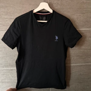 Svart t-shirt från U.S. Polo Assn. - Snygg svart t-shirt från U.S. Polo Assn. med en liten broderad logga på bröstet. T-shirten har en v-ringad hals och korta ärmar, perfekt för en stilren look.