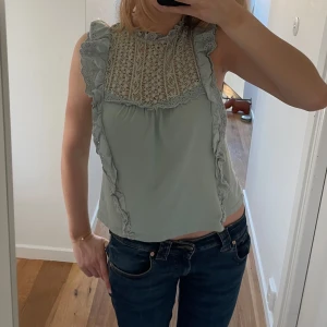 Grön ärmlös blus med spetsdetaljer - Snygg grön ärmlös blus med vackra spetsdetaljer och volanger. Perfekt för en somrig look. Blusen har en rund halsringning och en knapp i nacken.