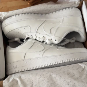 Vita Nike Air Force 1 - Klassiska vita Nike Air Force 1 sneakers med snörning och perforerade detaljer på tån. Skorna har en robust sula med Nike-logga och är tillverkade i skinn. Perfekta för en stilren look. Ny och är oanvänd 