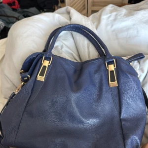 Blå handväska med gulddetaljer - Snygg blå handväska i skinn med eleganta gulddetaljer. Väskan har en praktisk dragkedja och rymligt inre med extra fack för småsaker. Perfekt för att bära med sig allt man behöver under dagen.