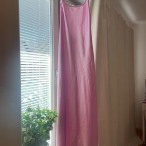 Rosa Satin långklänning - Säljer en elegant rosa långklänning med smala axelband. Klänningen har en enkel och stilren design, perfekt för somriga tillfällen. Materialet ser ut att vara lätt och luftigt, vilket gör den bekväm att bära. 